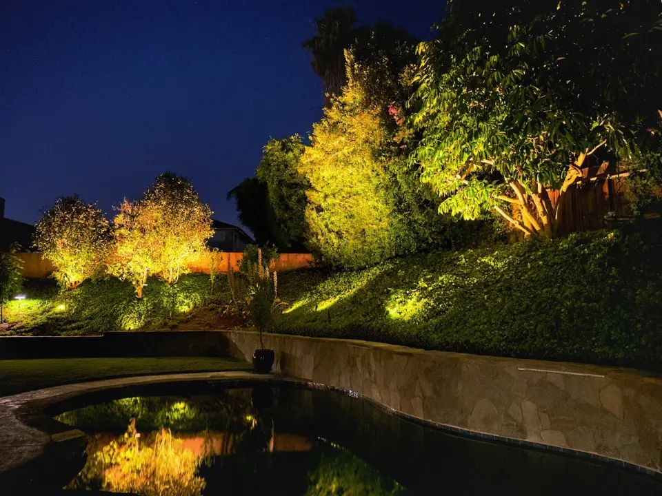 Tree and hedge uplighting installed along a sloped backyard near a pool in Laguna Hills, CA, highlighting mature trees and landscaping with warm golden light reflected in the water. Vision Outdoor Lighting designed and installed this landscape lighting to create depth and ambiance around the pool area at night. Vision Outdoor Lighting provides tree uplighting and pool-area landscape lighting throughout Laguna Hills and Orange County, California.