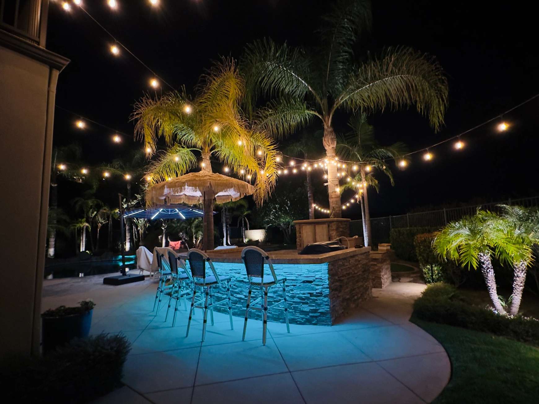 LED strip lighting installed under a stone outdoor bar, glowing blue, with string lights hanging through palm trees above a backyard pool in Coto de Caza, California at night.