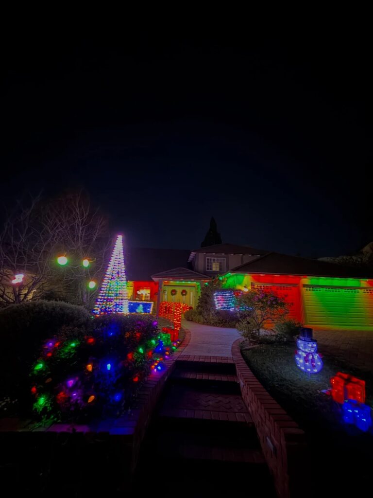 Two-story home in Anaheim Hills, CA decorated with a professional holiday and Christmas lighting installation, featuring a lit tree display, multicolor roofline and garage projections, wrapped garden bushes, and a lit snowman yard decoration along the front walkway.