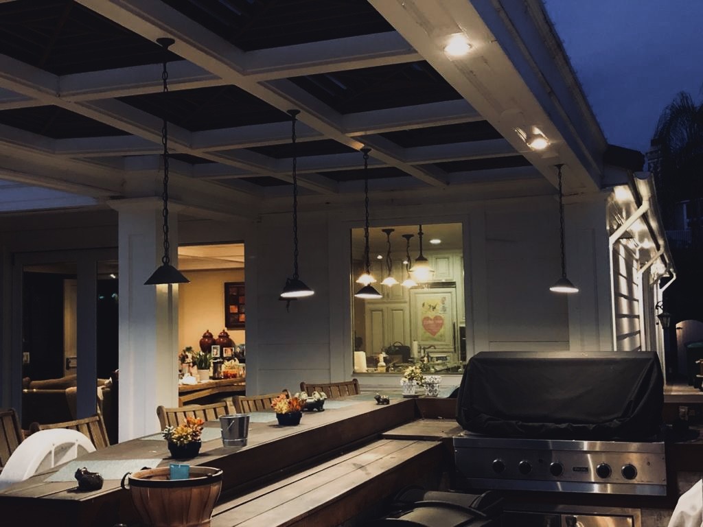 Covered outdoor kitchen with a built-in grill lit by hanging pendant lights along a coffered patio ceiling in the evening.
