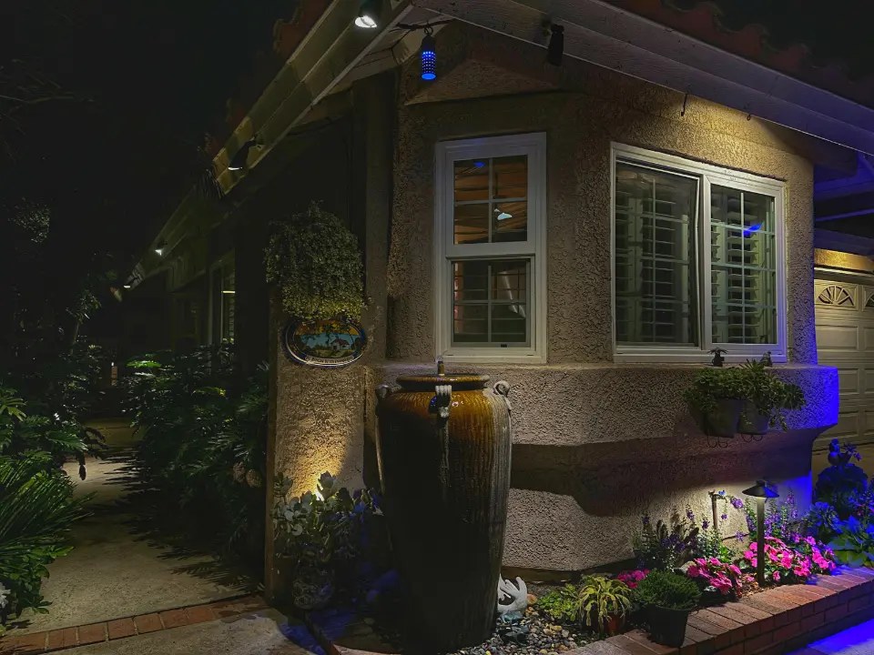 Ceramic urn wall fountain in Corona del Mar, CA illuminated with warm accent lighting and a blue color-changing wash across the adjacent flower bed and garage wall at night.