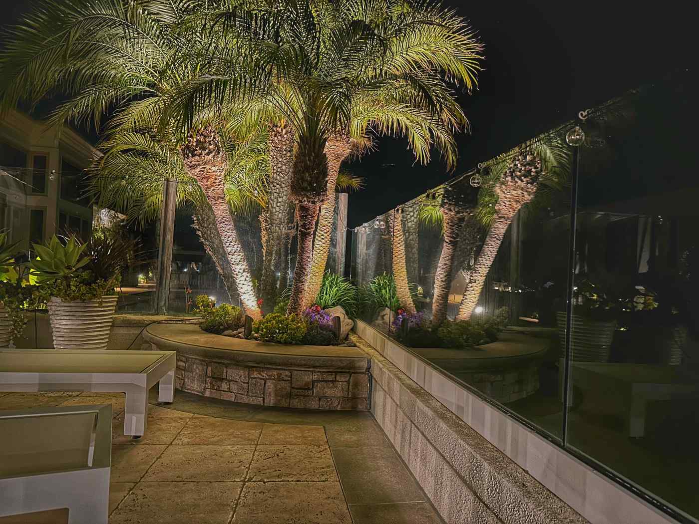 Cluster of palm trees in a raised stone planter uplit at night beside a glass-railed patio seating area.