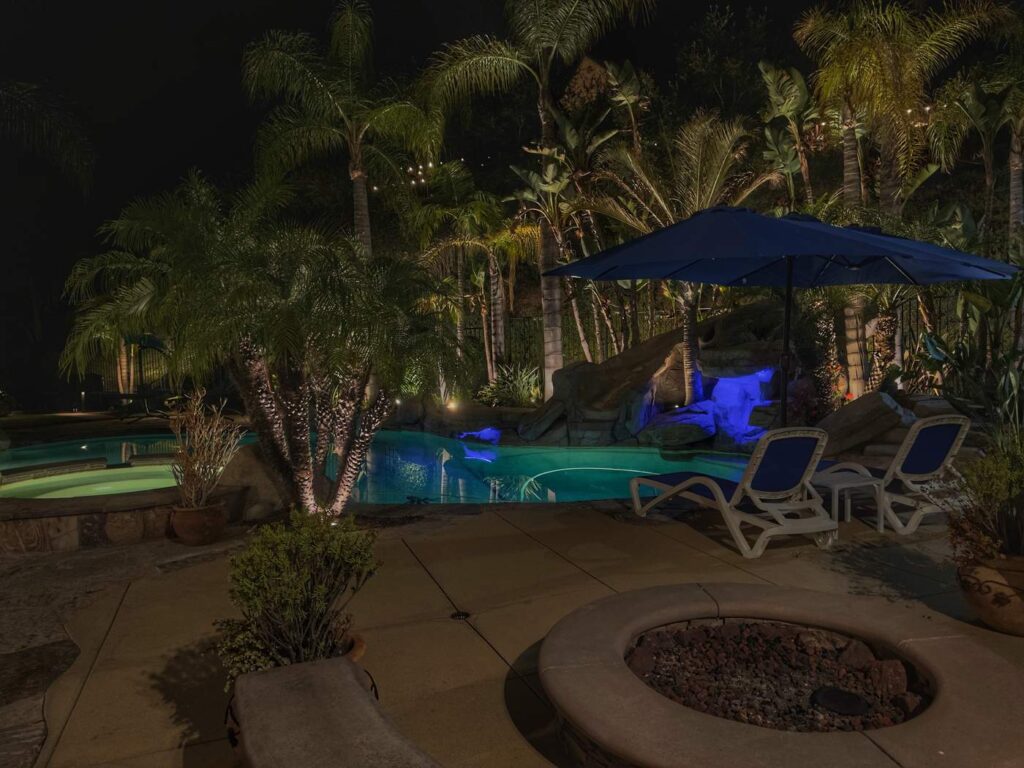 Backyard pool and spa in Coto de Caza, CA with a rock waterslide accented by blue color-changing lighting, framed by uplit palm trees and a shaded lounge area at night.