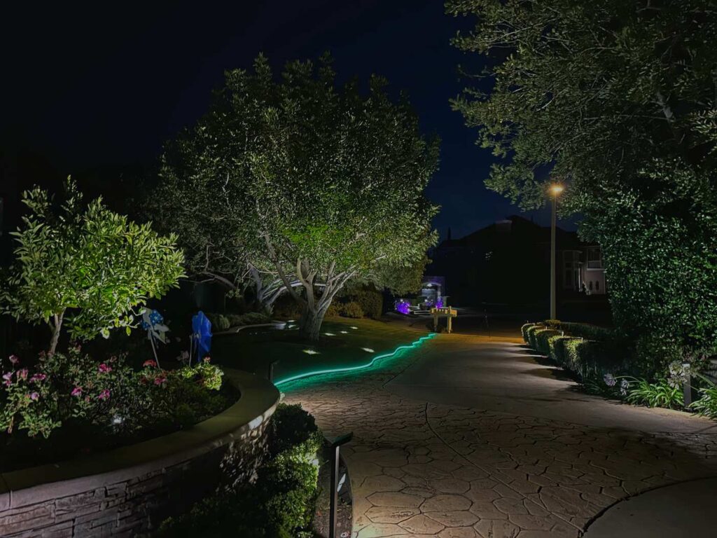 Stamped concrete driveway edge in Coto de Caza, CA outlined with a green color-changing LED strip light winding along the stonework toward the front entry at night.