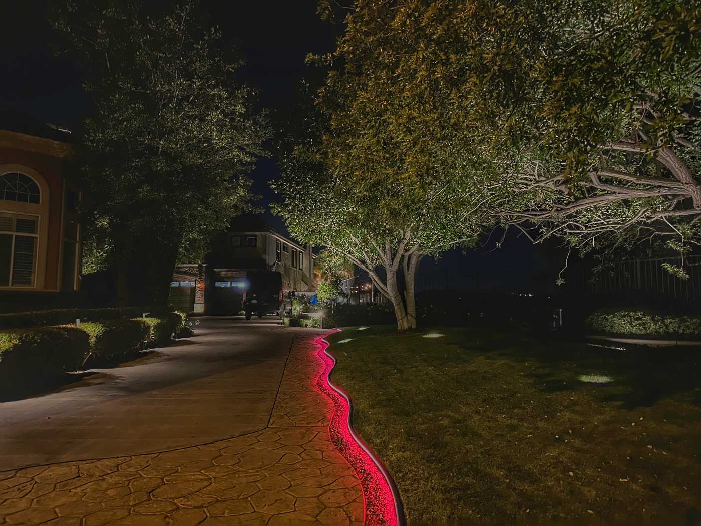 Stamped concrete driveway edge in Coto de Caza, CA outlined with a red color-changing LED strip light winding past uplit trees toward a home entry at night.