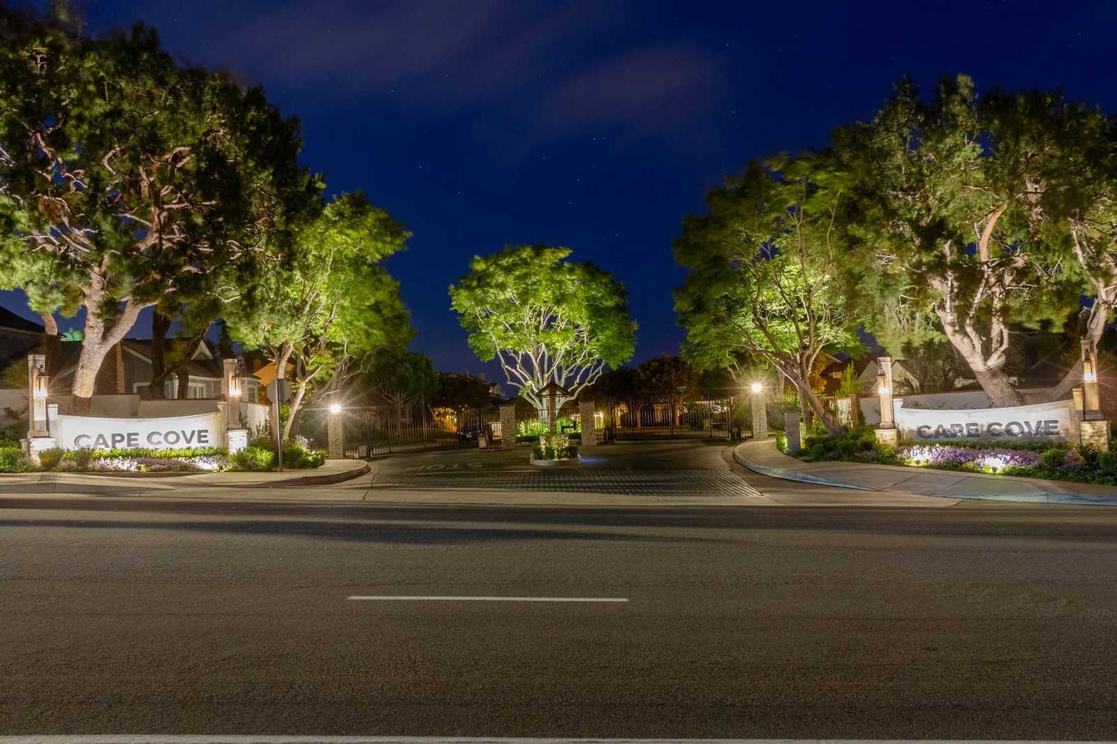 Mature trees are uplit at the gated entrance of the Cape Cove community, framing the illuminated stone entry signage and wrought iron gates at night.