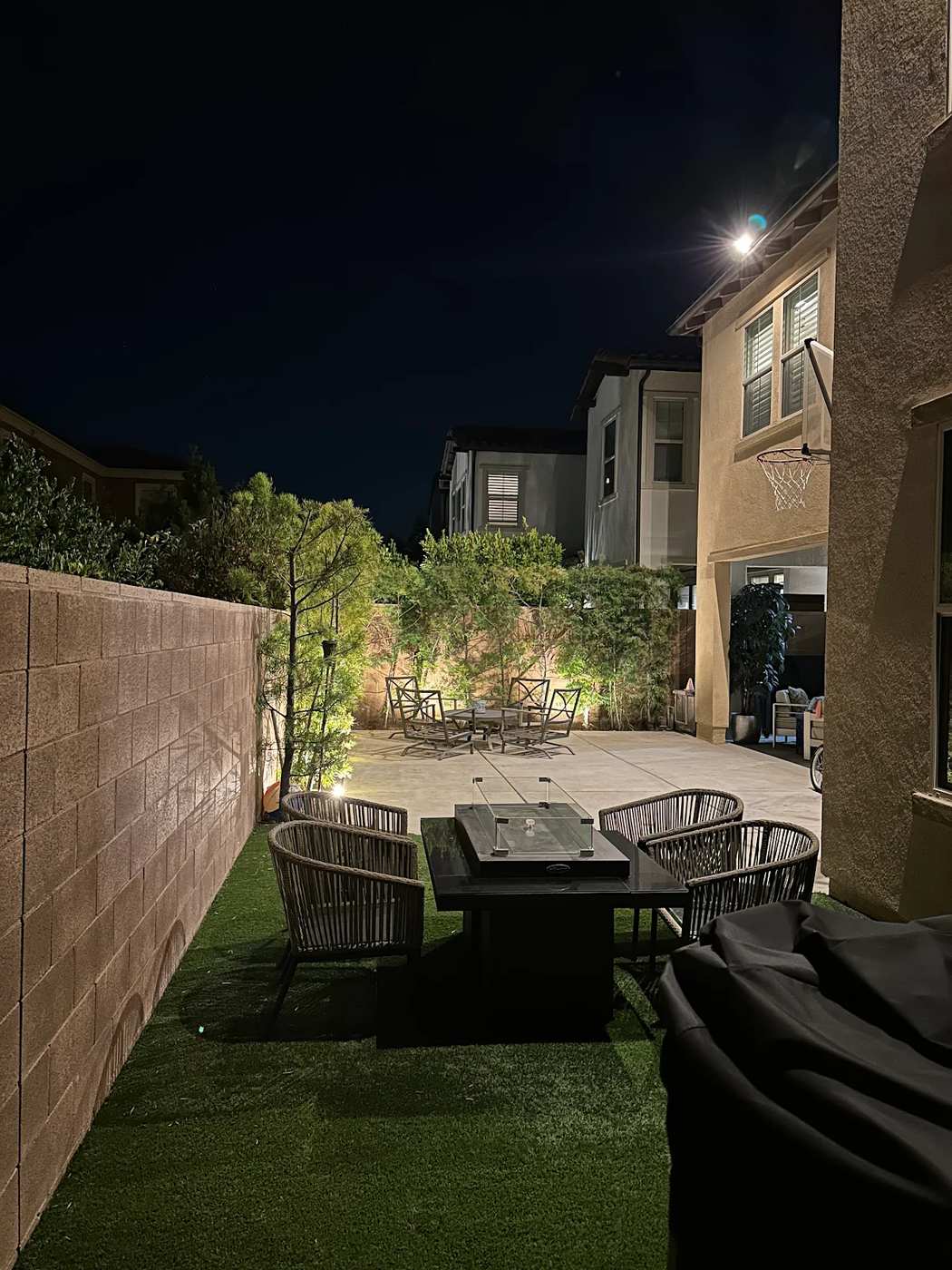 Backyard patio in Fullerton, CA with artificial turf and a seating area beside an uplit block wall and ornamental tree, with a covered outdoor dining space visible beyond at night.