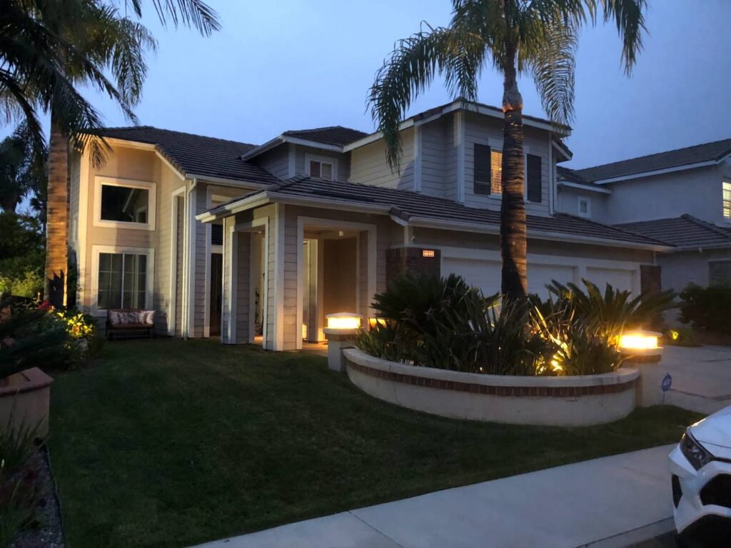 Two-story home in Fullerton, CA at dusk with illuminated post lights atop a curved brick planter wall, accenting palm trees and tropical foliage along the front entry path.