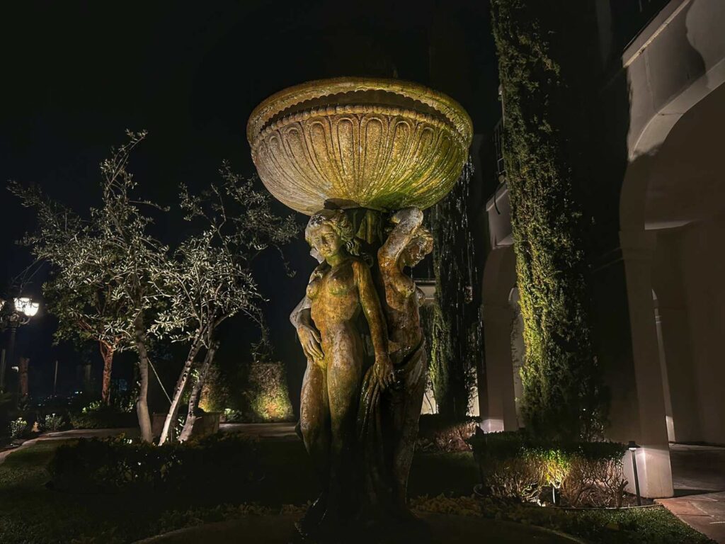A tiered stone fountain with sculpted figures is lit at night alongside uplit olive trees and an ivy-covered wall, creating a dramatic garden focal point.