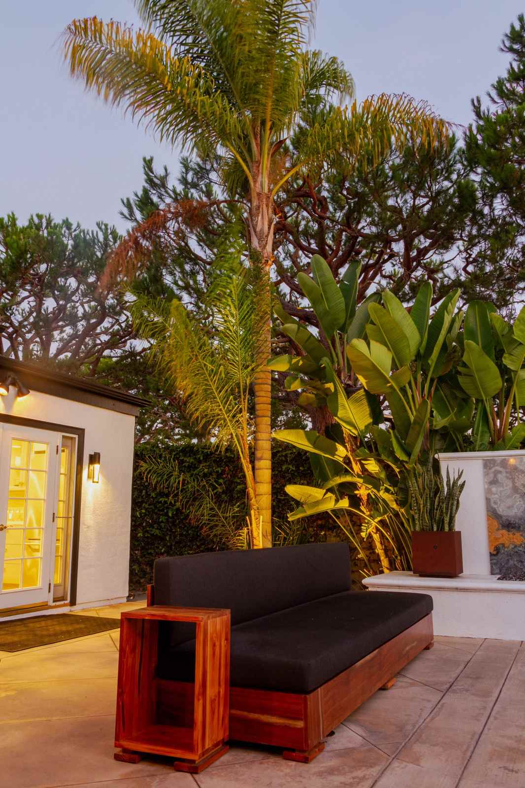 A tall palm and bird of paradise plants are uplit against a backyard patio with a built-in sofa, string lights, and a lit guest house in the background at dusk.