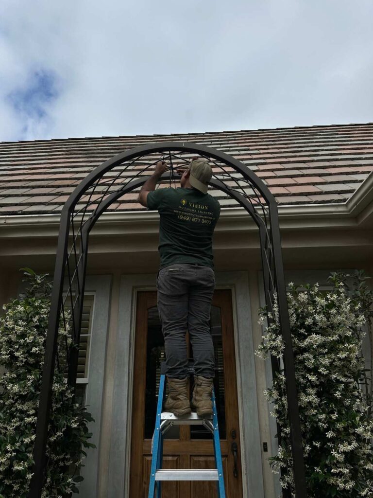 A Vision Outdoor Lighting technician installs string lighting along an iron arch pergola over a jasmine-covered front entryway.