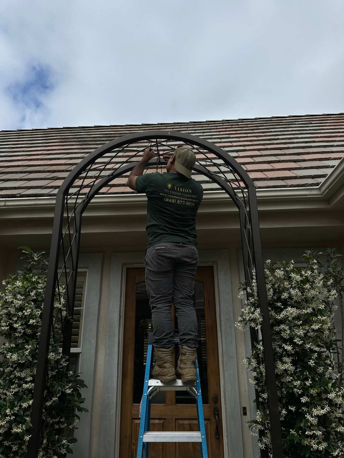 A Vision Outdoor Lighting technician installs string lighting along an iron arch pergola over a jasmine-covered front entryway.