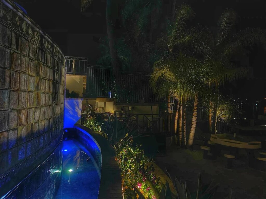 A curved stone wall and lap pool are lit in blue accent lighting alongside uplit palm trees and a patio dining area at night.