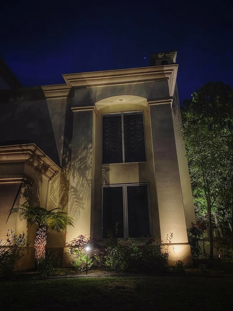 A tall stucco estate facade with an arched window is uplit at night, with a fern and shrub bed lit along the base of the wall.