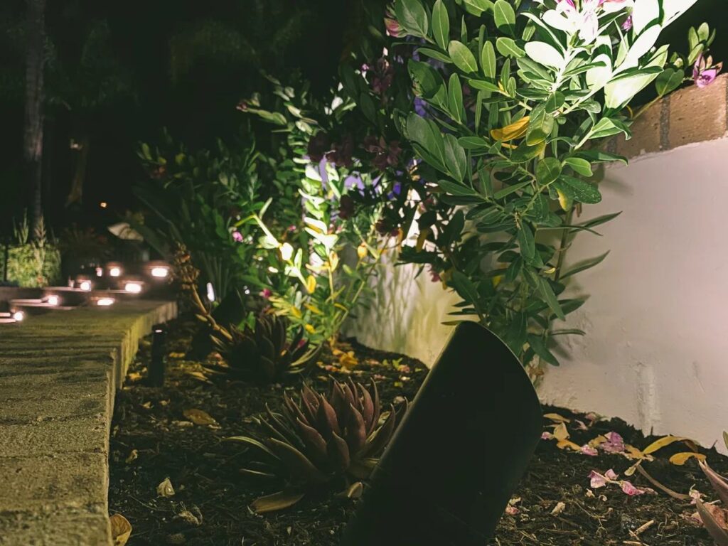 Path lights and uplighting accent a succulent and flowering garden bed along a stepped stone walkway beside a stucco wall.