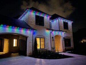 Two-story stone home in Irvine, CA featuring a permanent color-changing outdoor lighting system installed along the roofline and eaves by Vision Outdoor Lighting, shown illuminated at night in a rainbow color pattern.