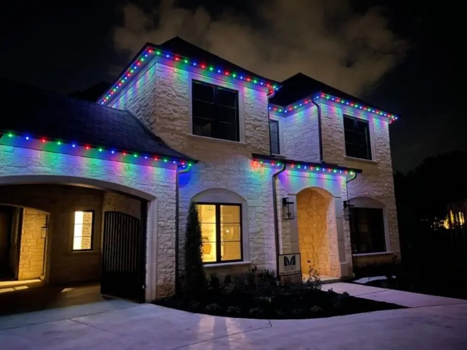 Two-story stone home in Irvine, CA featuring a permanent color-changing outdoor lighting system installed along the roofline and eaves by Vision Outdoor Lighting, shown illuminated at night in a rainbow color pattern.