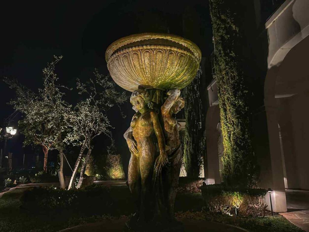 A large weathered stone fountain supported by classical figures lit from below in a garden courtyard at night, with uplit Italian cypress trees and an olive tree behind it.