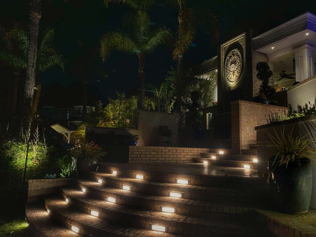 Recessed step lights illuminate tiered brick staircases leading to a home entry with a decorative medallion wall and lit palm trees.