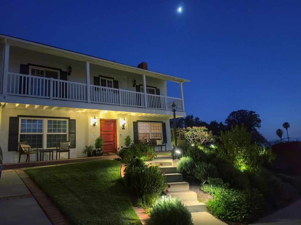 Path lights and garden uplights illuminate the front lawn and steps of a two-story colonial home with a wrap-around porch at dusk.