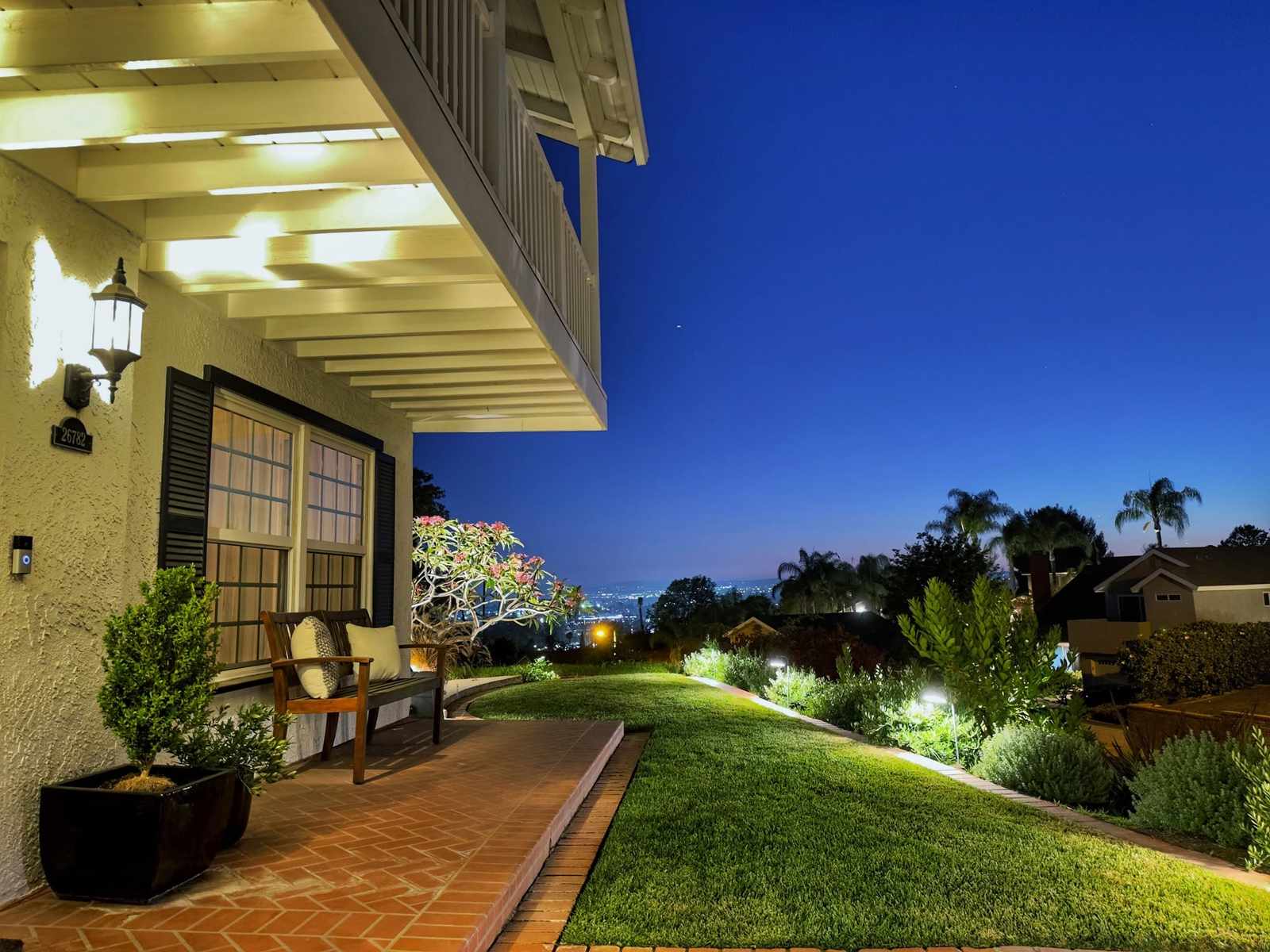 Garden path lights illuminate a sloped front lawn and porch area of a colonial home at dusk, with city lights and palm trees visible in the distance.