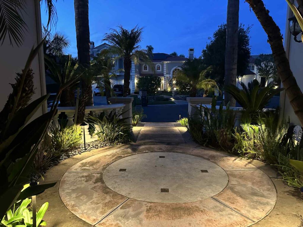 Circuxlar stone driveway entrance in Santa Ana, CA framed by uplit palm trees, sago palms, and garden bed foliage along a stone balustrade, with a two-story home glowing softly in the background at dusk.