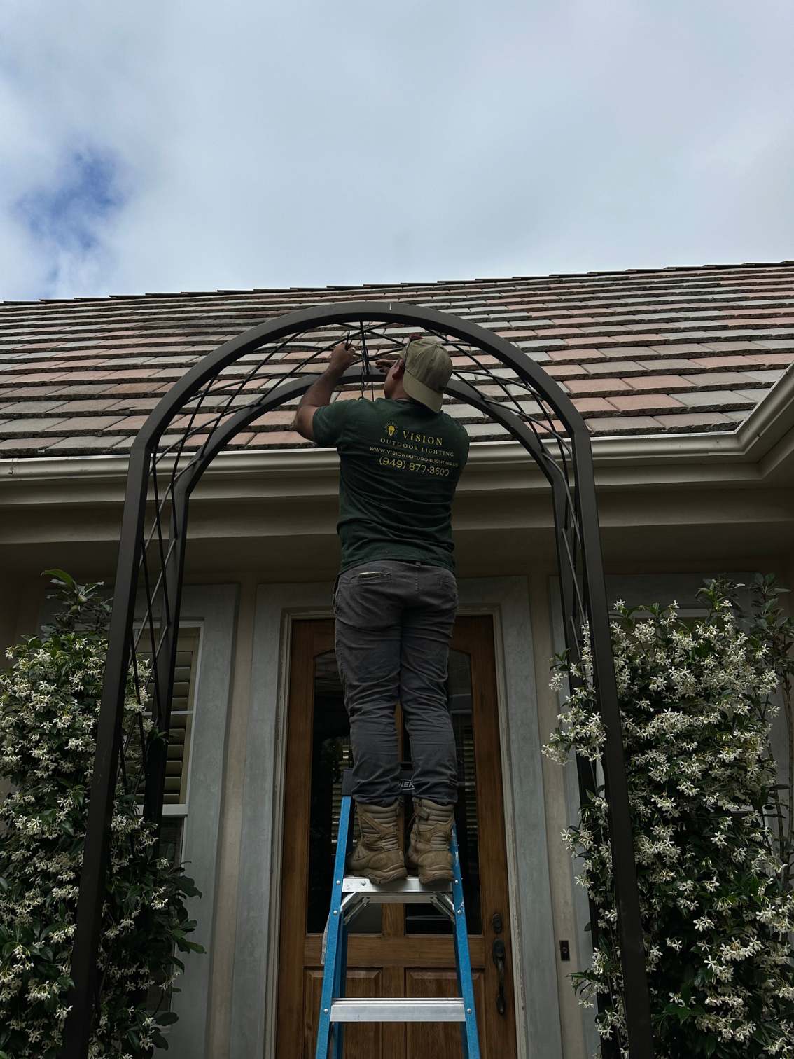 Vision Outdoor Lighting technician installing accent lighting along an iron garden arch entryway in Santa Ana, CA, working from a ladder amid climbing star jasmine vines in front of a tile-roofed home.