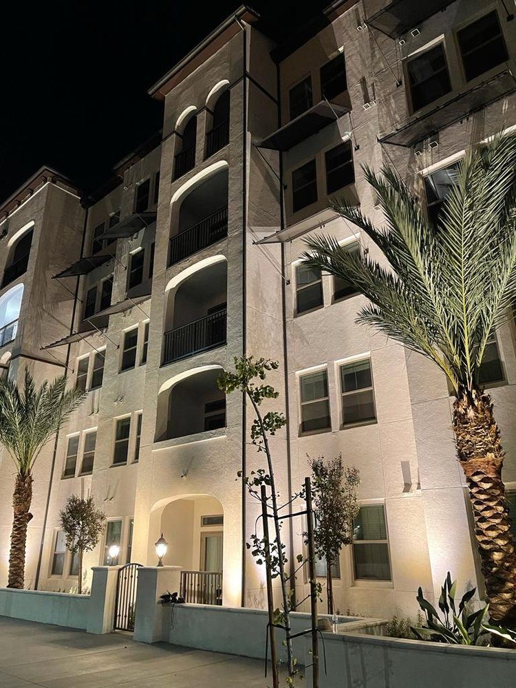 Commercial building facade uplighting on a Mediterranean-style multi-story property in Orange County CA with arched balcony illumination and palm tree accent lighting at night