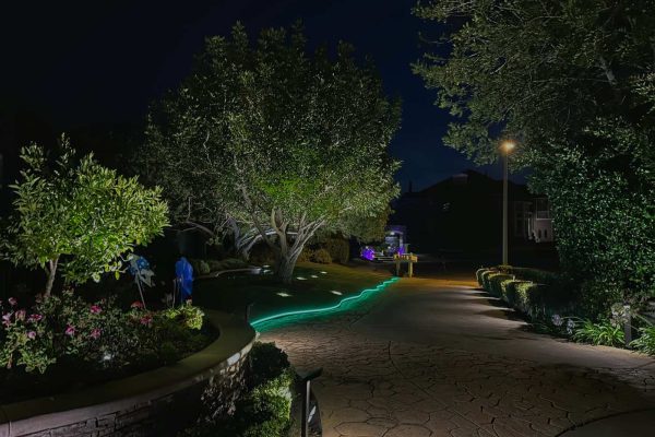 Stamped concrete driveway edge in Coto de Caza, CA outlined with a green color-changing LED strip light winding along the stonework toward the front entry at night.