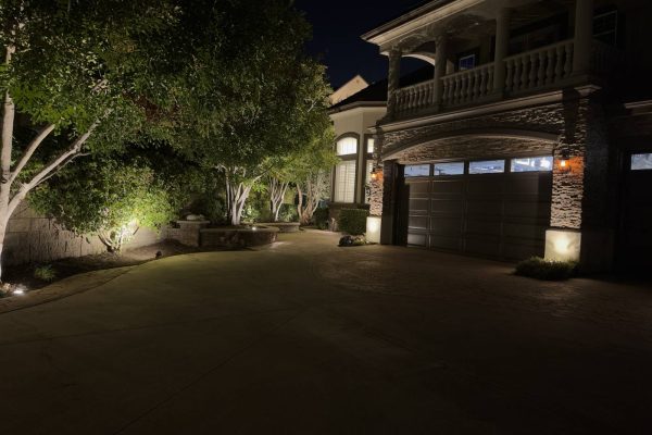 Circular driveway in Coto de Caza, CA leading to a stone-accented garage, with uplit ornamental trees and low path lights lining the planter beds at night.