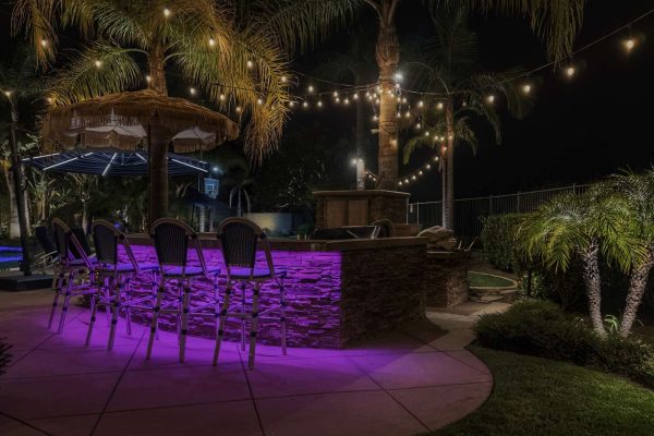 Outdoor stone bar in Coto de Caza, CA lit with purple LED strip lighting beneath the counter, surrounded by string lights strung through palm trees and a thatched-roof umbrella at night.