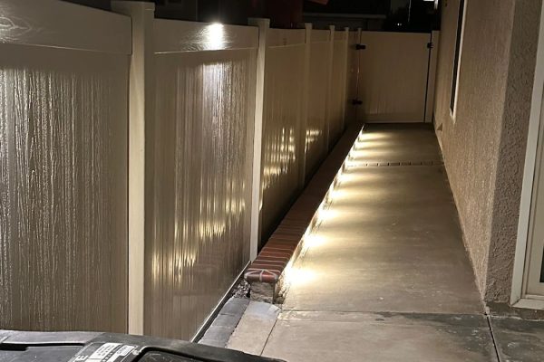 Side yard walkway in Fullerton, CA lined with low path lights along a brick border and vinyl fence, leading toward a rear gate at night.