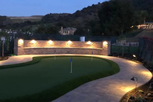 A backyard putting green is framed by an uplit stacked-stone accent wall at dusk, with rolling hillside views in the background.
