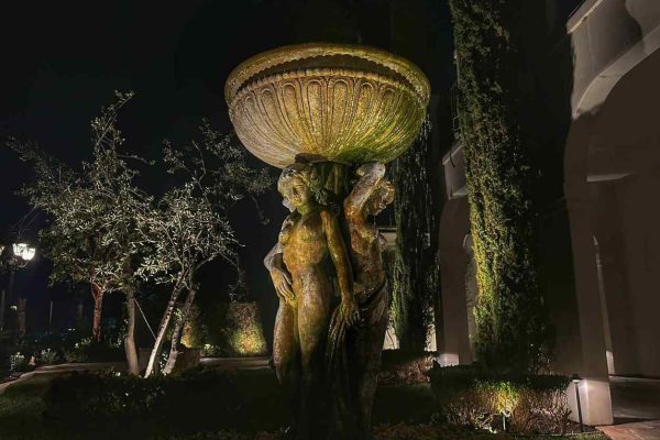 A large weathered stone fountain supported by classical figures lit from below in a garden courtyard at night, with uplit Italian cypress trees and an olive tree behind it.