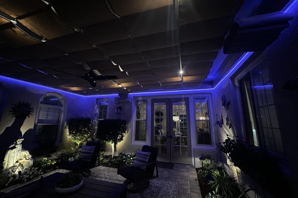 Covered patio in Tustin, CA with a retractable canvas shade ceiling outlined by a blue color-changing LED strip light along the perimeter, illuminating garden statuary and potted trees at night.